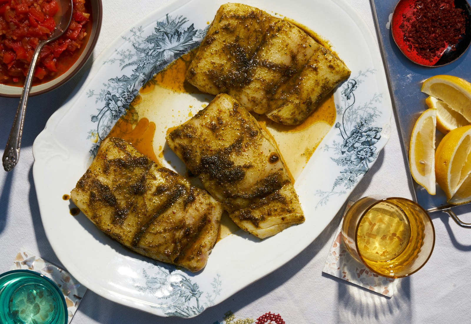 Sea bass with chermoula on grey and white plate alongside lemon wedges and bowl of chopped tomatoes.