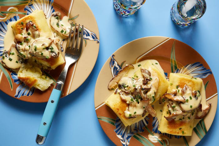 Savory blintzes with creamy mushroom sauce and chives on colorful plates atop blue surface.