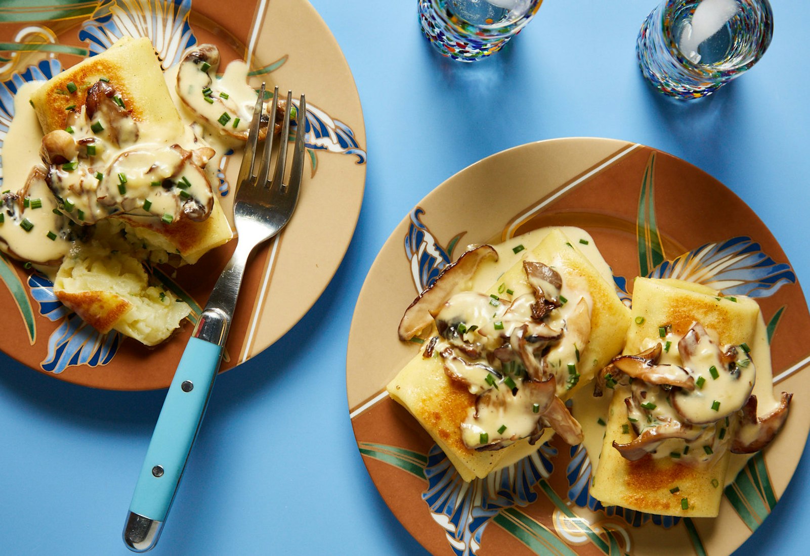 Savory blintzes with creamy mushroom sauce and chives on colorful plates atop blue surface.
