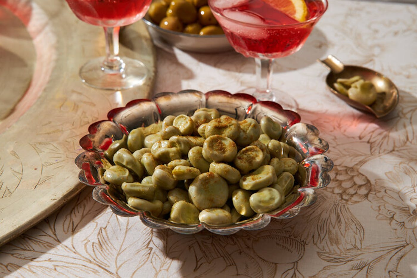 Fava beans sprinkled with cumin, small bowl for peels, pink cocktail with lemon.