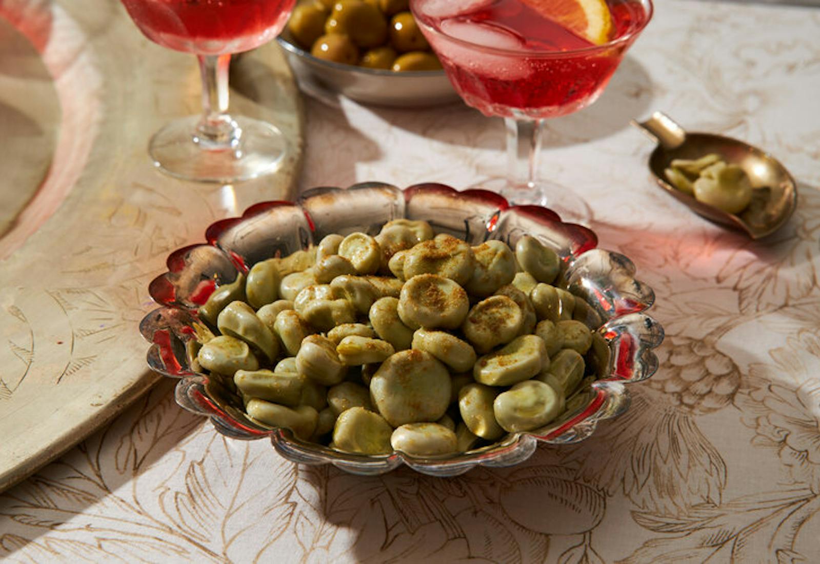 Fava beans sprinkled with cumin, small bowl for peels, pink cocktail with lemon.