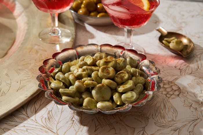 Fava beans sprinkled with cumin, small bowl for peels, pink cocktail with lemon.