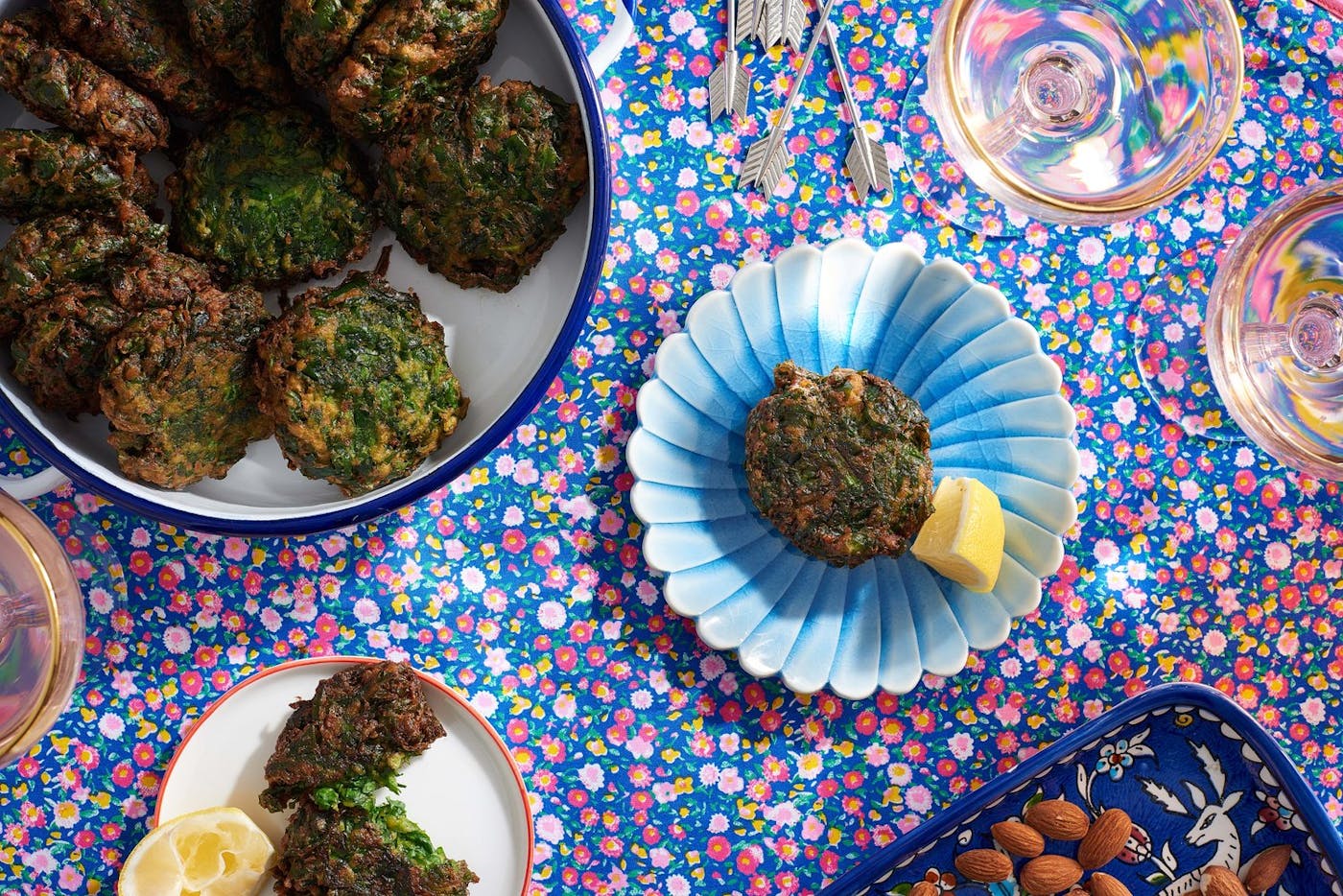 Servings of spinach rissoles with lemon wedges alongside large dish of rissoles, dish with almonds and glasses of white wine atop blue and pink floral tablecloth.