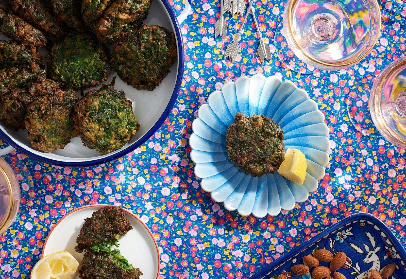 Servings of spinach rissoles with lemon wedges alongside large dish of rissoles, dish with almonds and glasses of white wine atop blue and pink floral tablecloth.