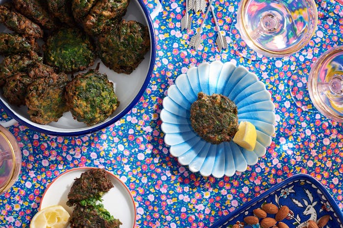 Servings of spinach rissoles with lemon wedges alongside large dish of rissoles, dish with almonds and glasses of white wine atop blue and pink floral tablecloth.