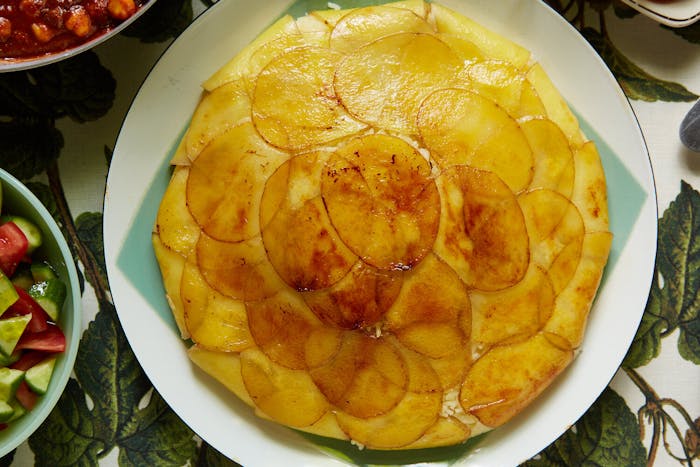 Potato tahdig on blue and white plate alongside cucumber and tomato salad, atop leaf-printed tablecloth.