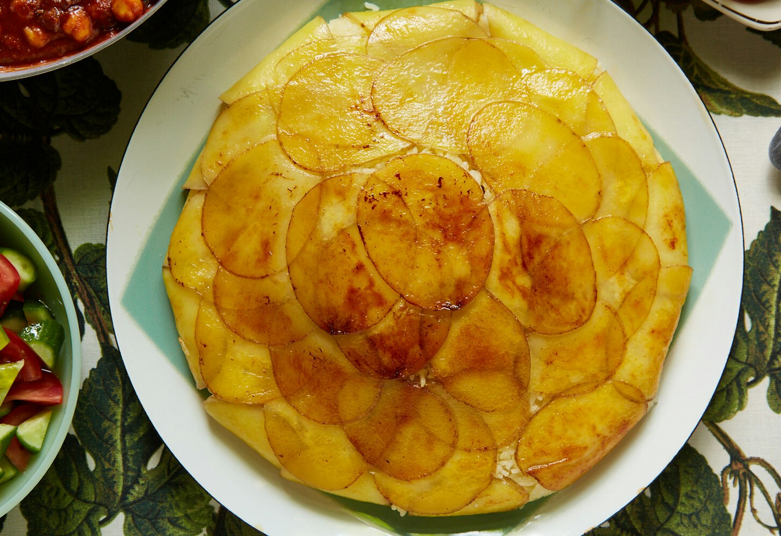 Potato tahdig on blue and white plate alongside cucumber and tomato salad, atop leaf-printed tablecloth.