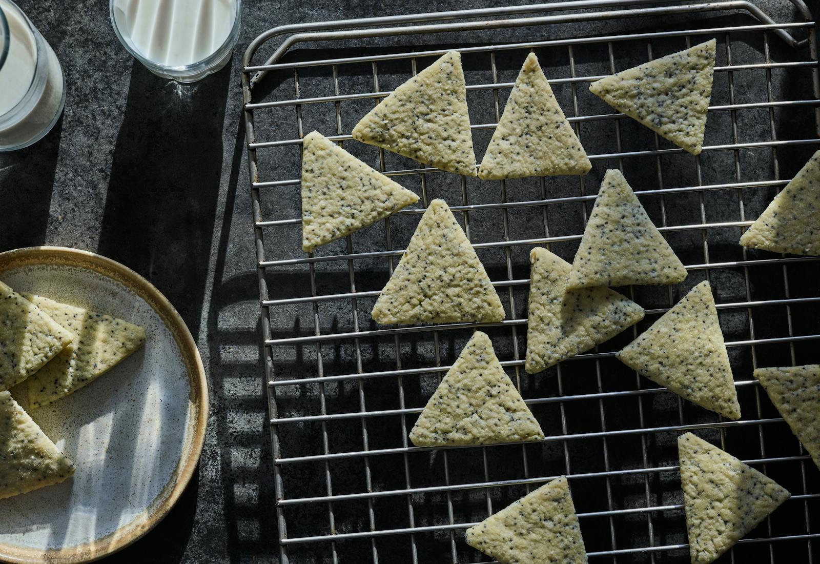 Triangular poppyseed cookies on cooling rack with a glass of milk beside them.