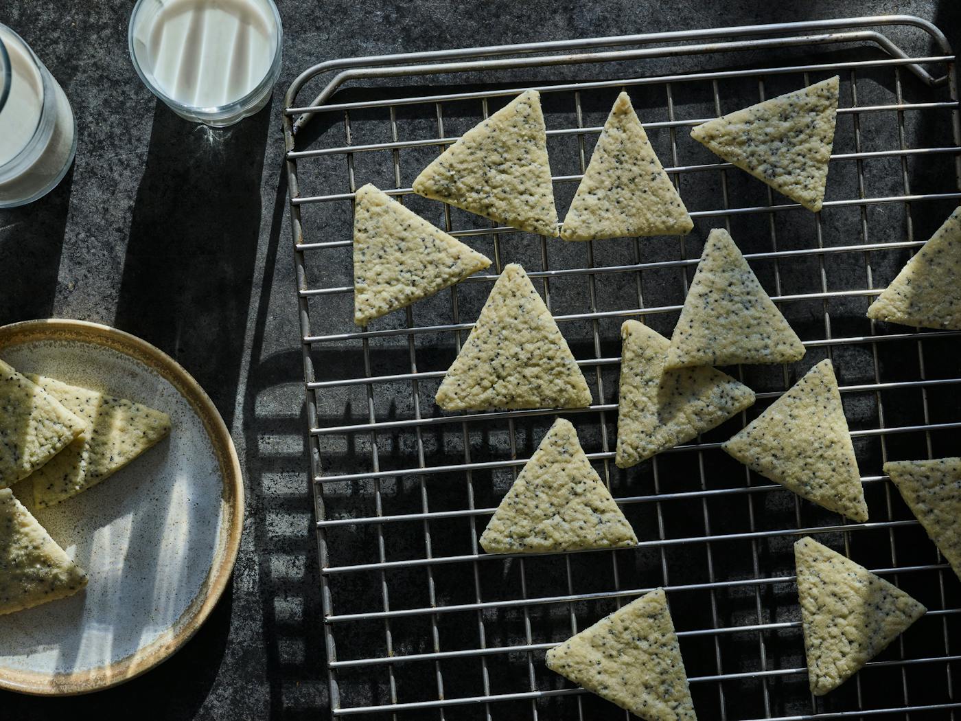 Triangular poppyseed cookies on cooling rack with a glass of milk beside them.