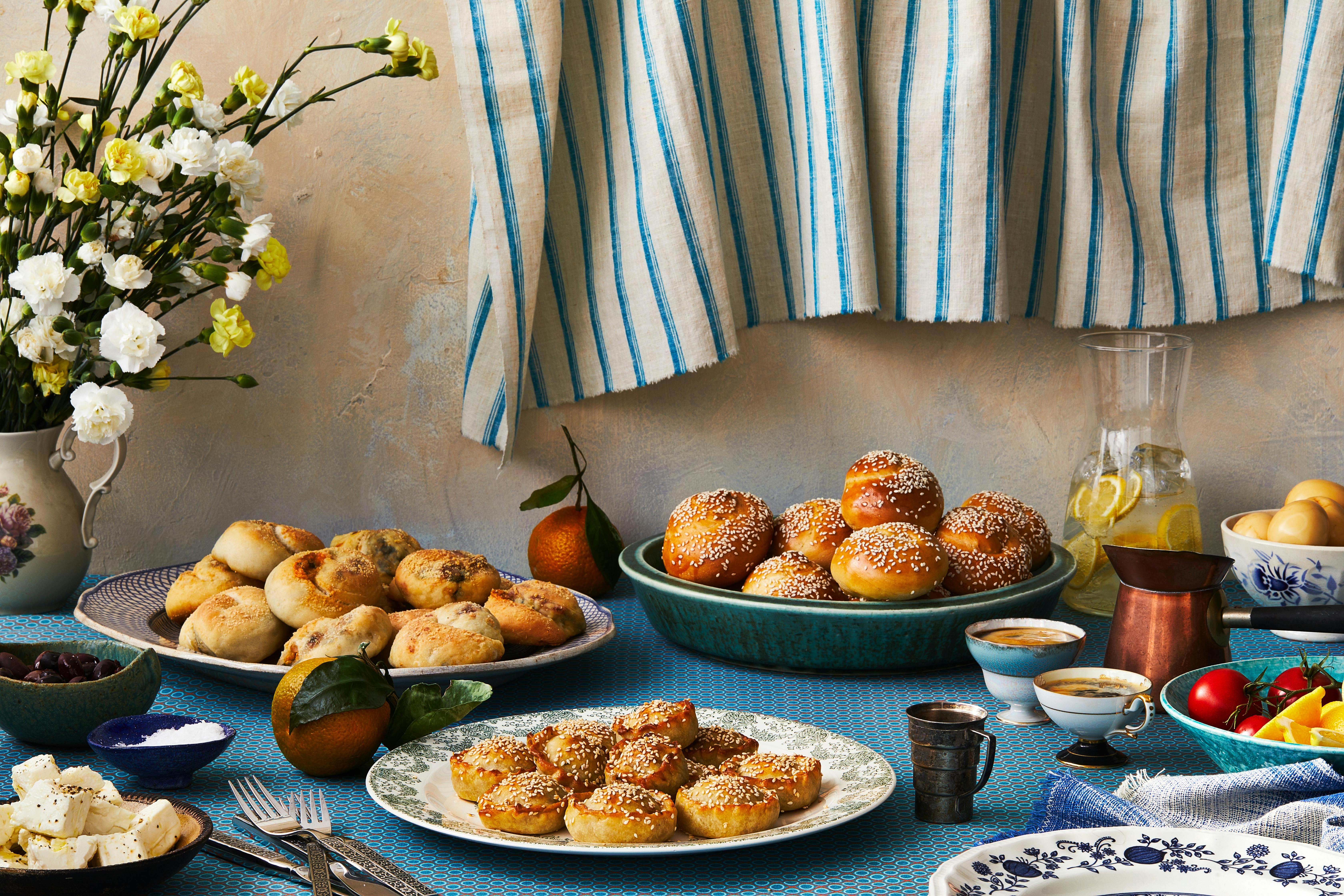 Capeluoto table spread of plate of boyos, pastelikos, and roskas over blue tablecloth with fruits and coffee on the side..