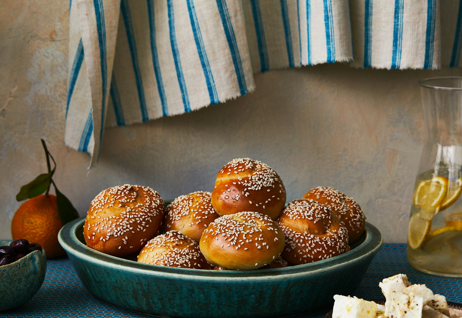 Roskas stacked in teal bowl atop blue tablecloth, alongside pitcher of lemon water.