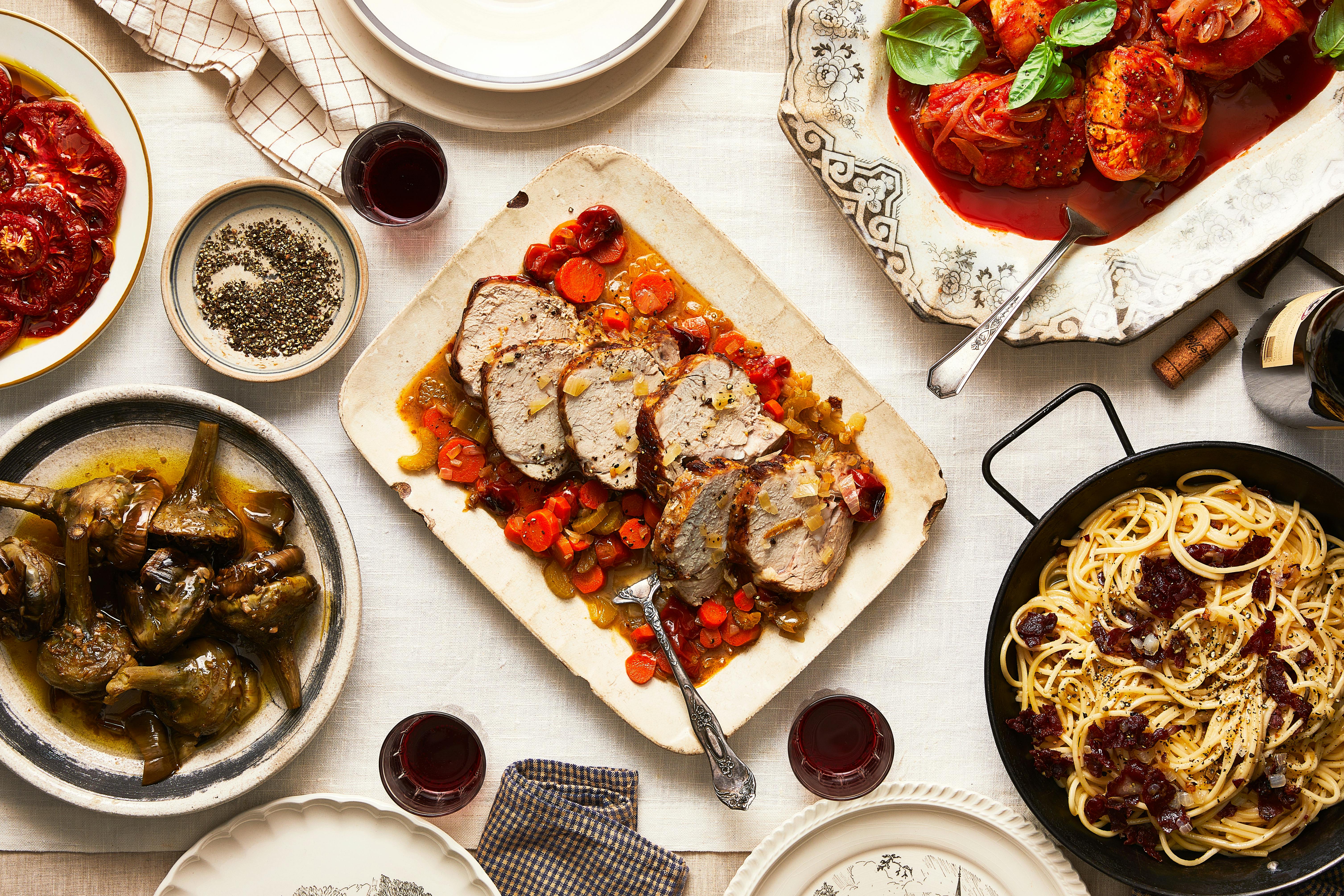 Sara's Roman Jewish table spread with vitella al forno in the center surrounded by Roman carbonara, Roman-style artichokes, baccala with onion and tomato, and pomodori a mezzo, over a white tablecloth.