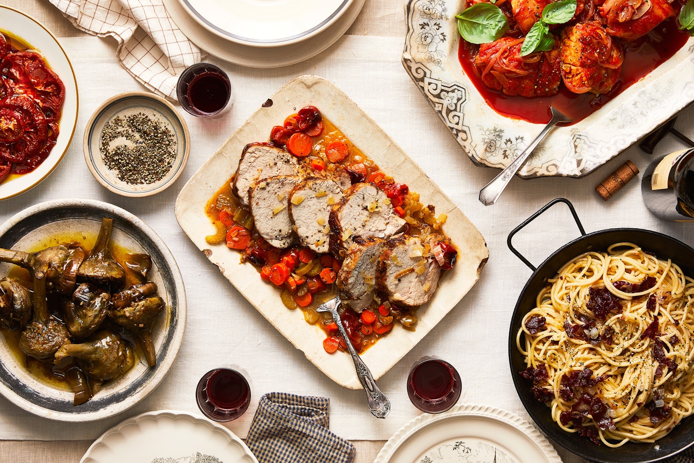 Sara's Roman Jewish table spread with vitella al forno in the center surrounded by Roman carbonara, Roman-style artichokes, baccala with onion and tomato, and pomodori a mezzo, over a white tablecloth.