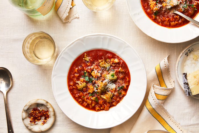 Broccoli soup with pasta and anchovy in white serving bowl over white linen tablecloth with a cloth napkin, cup of white wine and pecorino cheese on the side.