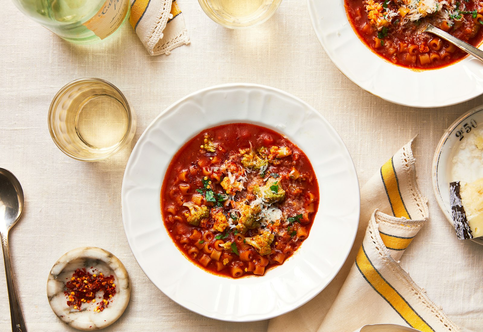 Broccoli soup with pasta and anchovy in white serving bowl over white linen tablecloth with a cloth napkin, cup of white wine and pecorino cheese on the side.