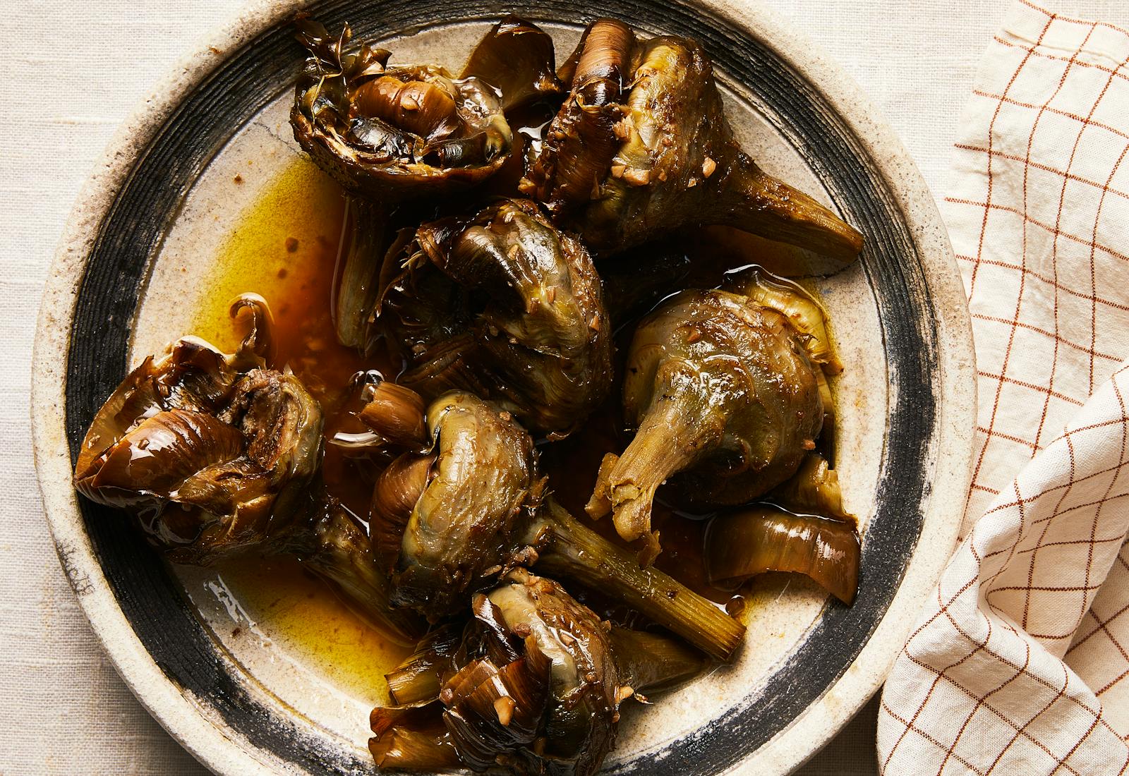 Braised Roman artichokes on beige serving plate with black rim atop white linen tablecloth.