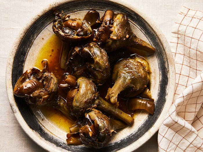 Braised Roman artichokes on beige serving plate with black rim atop white linen tablecloth.