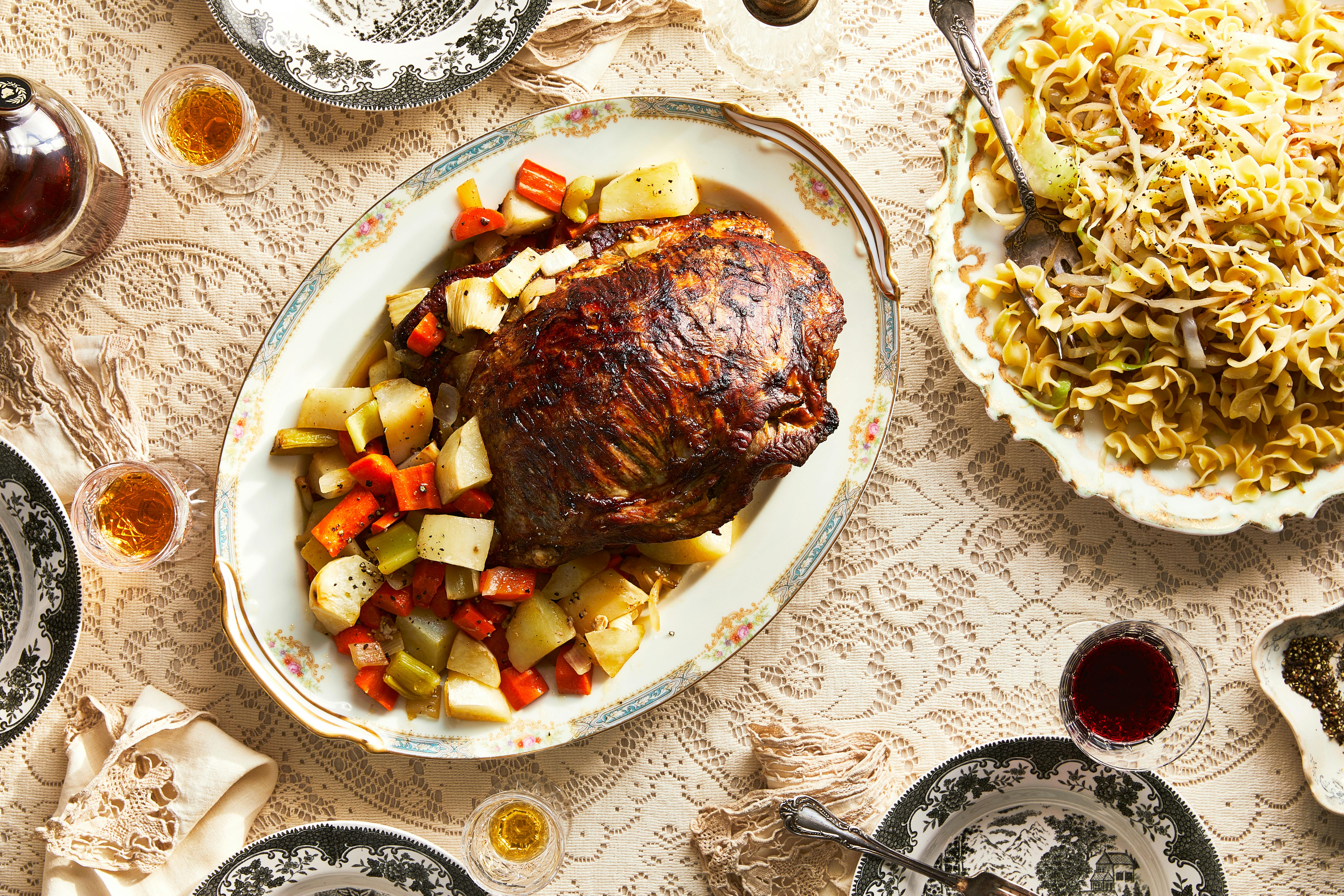 A Slovakian table set with a stuffed veal breast with roasted vegetables, sweet cabbage noodles, and glasses of wine alongside, atop a cream-colored lace tablecloth.