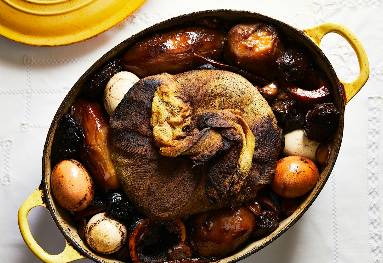 Large yellow Dutch oven filled with hamin: Russet potatoes, hard-boiled eggs, dates, beef marrow bones, wheat berries wrapped in cheesecloth and beef brisket, atop white tablecloth.