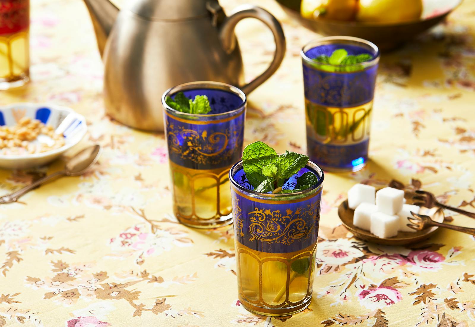 3 cups of mint tea garnished with fresh mint, sugar cubes on the side, silver teapot in the background, served over yellow floral tablecloth.