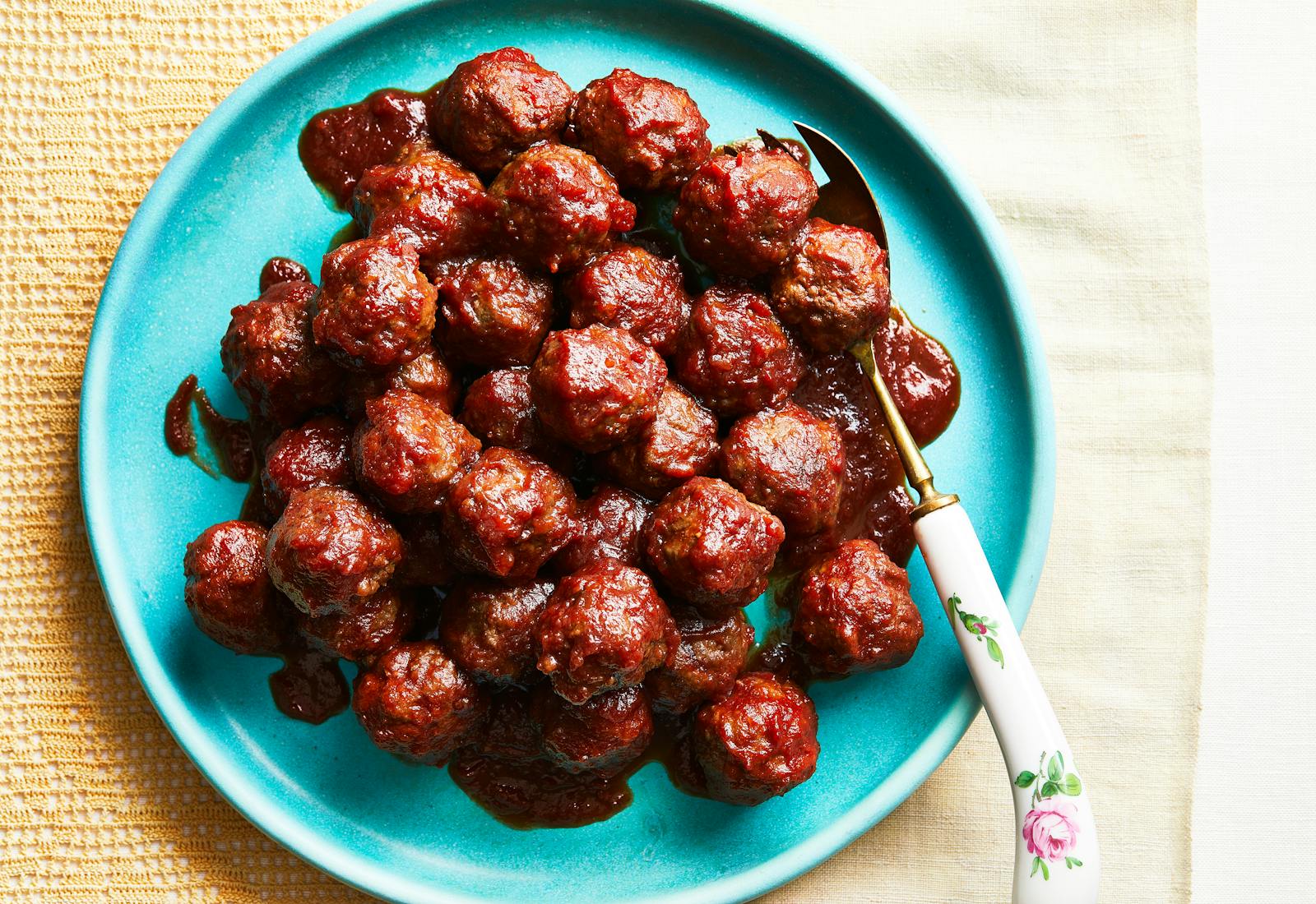 Meatballs stacked on a turquoise plate with a serving fork on the side atop yellow tablecloth.