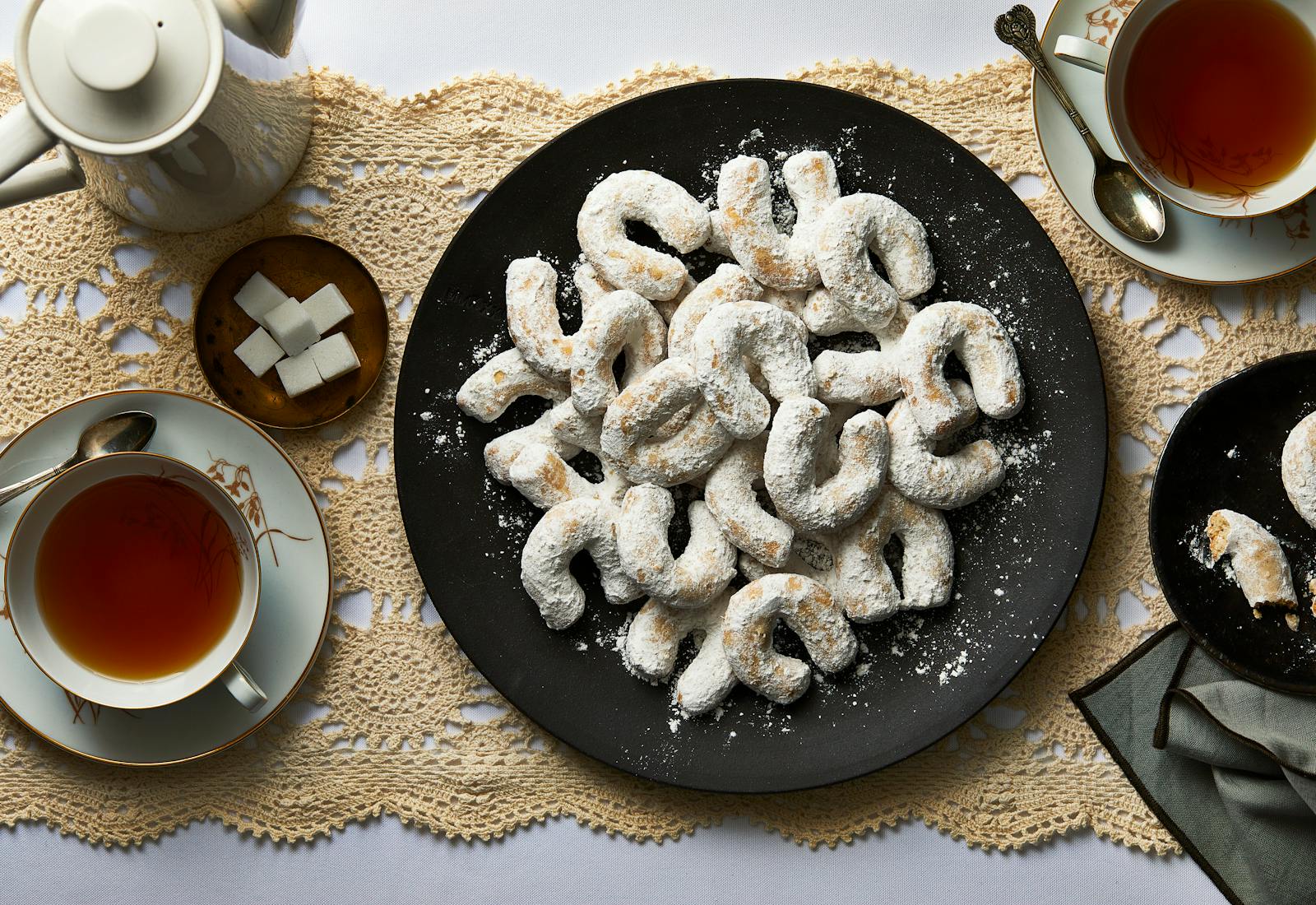 Plate of vanillekipferl served on black plate with tea and sugar on the side, atop beige latice table runner.