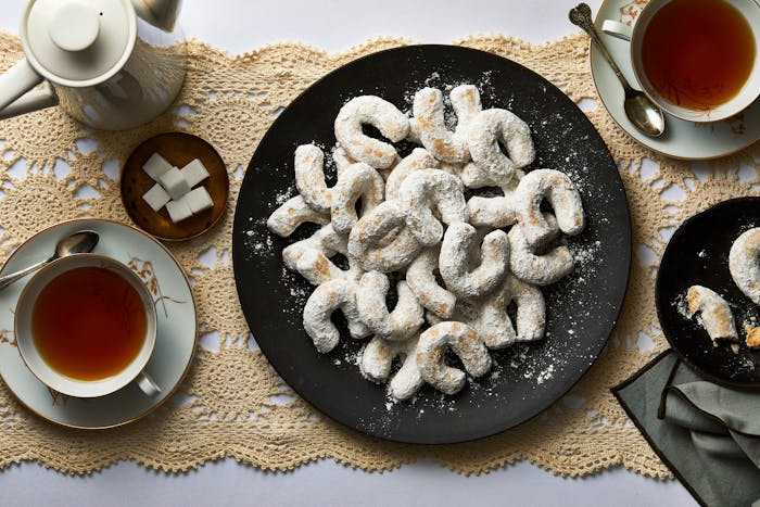 Plate of vanillekipferl served on black plate with tea and sugar on the side, atop beige latice table runner.