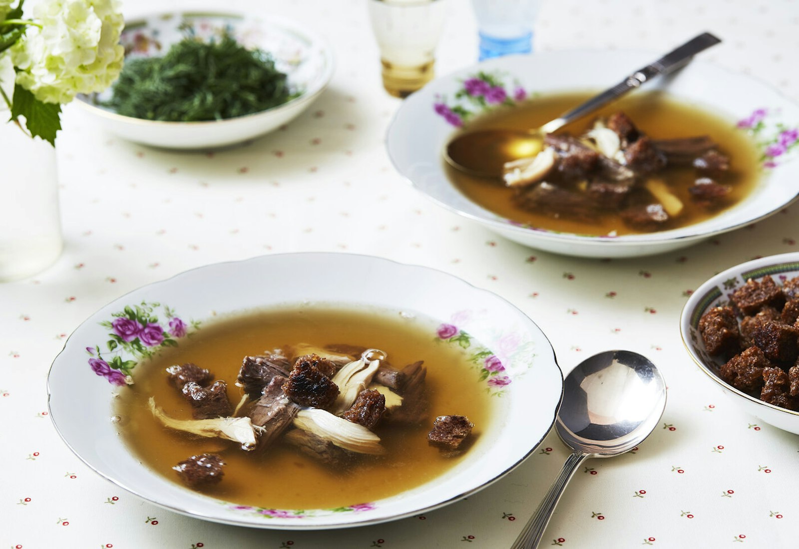 Bouillon in purple floral bowl, fresh flowers, bowl of croutons.