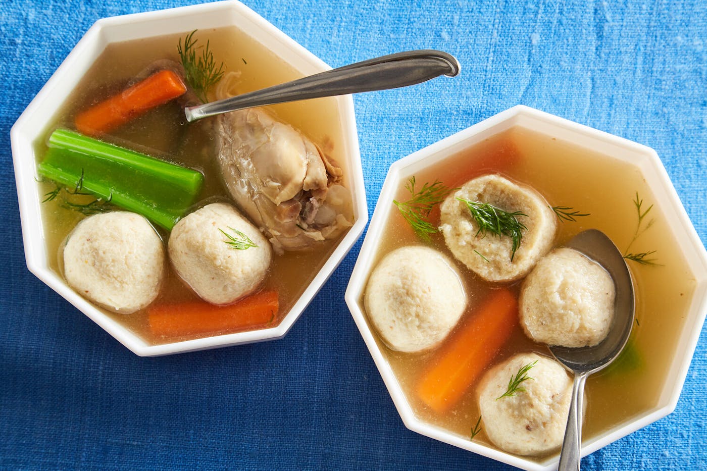 Matzo ball soup sprinkled with dill in two geometric bowls atop blue tablecloth.
