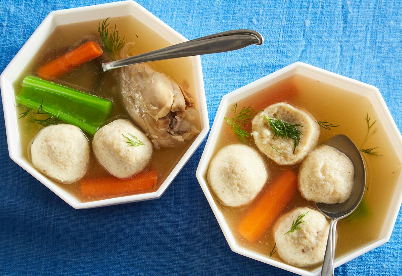 Matzo ball soup sprinkled with dill in two geometric bowls atop blue tablecloth.