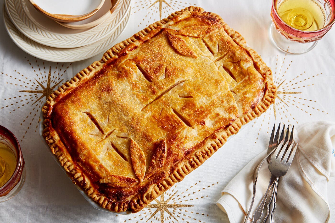 Apple strudel alongside glass of white wine and serving forks and plates, atop white tablecloth with star patterns.