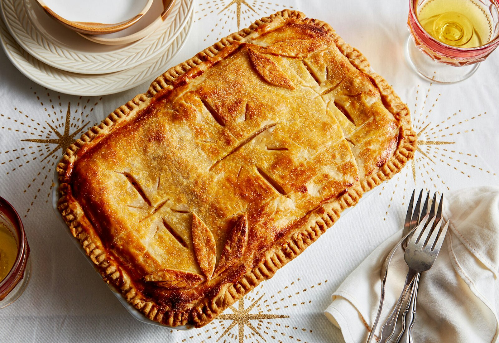 Apple strudel alongside glass of white wine and serving forks and plates, atop white tablecloth with star patterns.