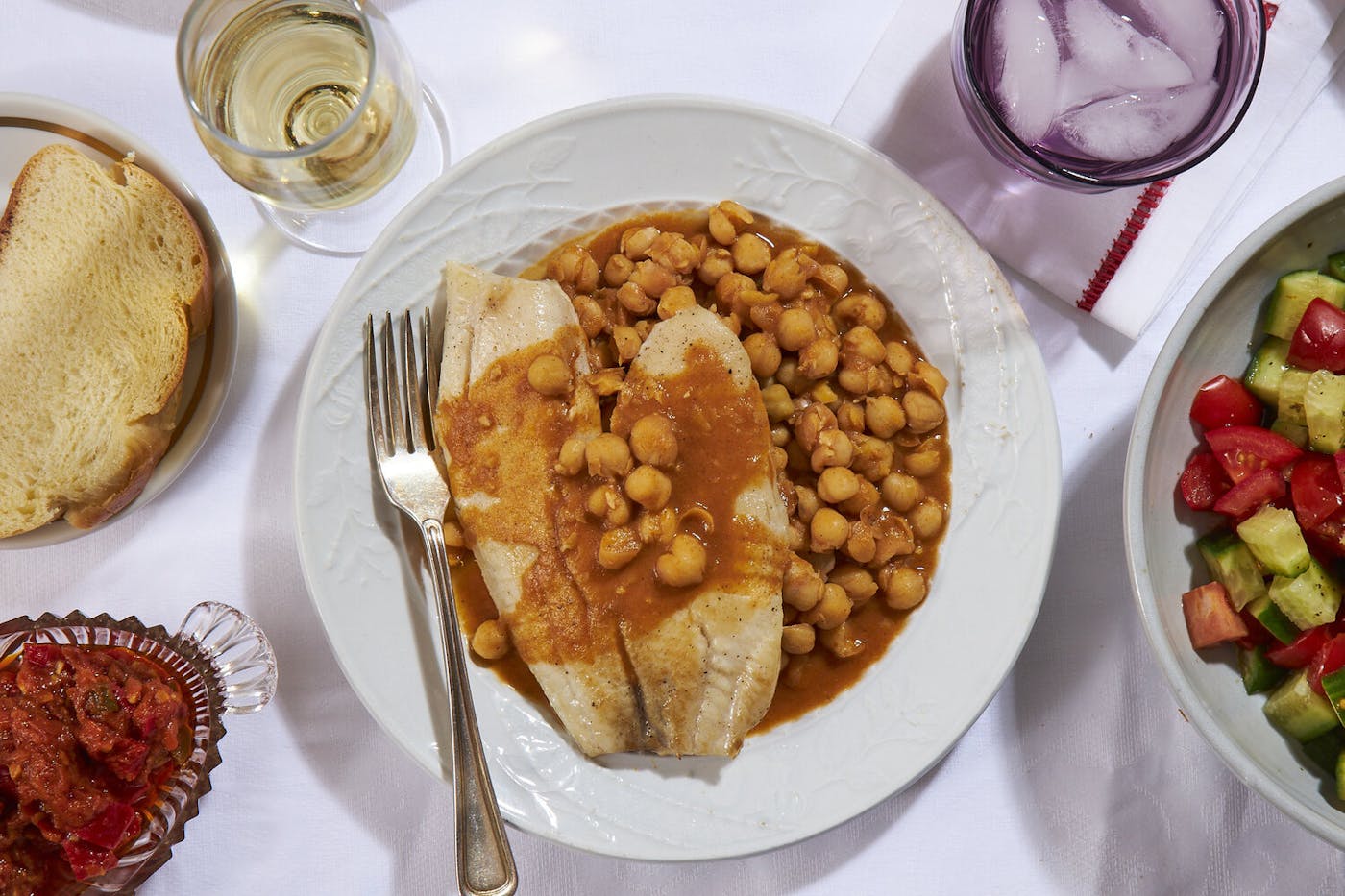 Chickpea stew with fish, slice of challah, white wine, matbucha and chopped salad atop white tablecloth.