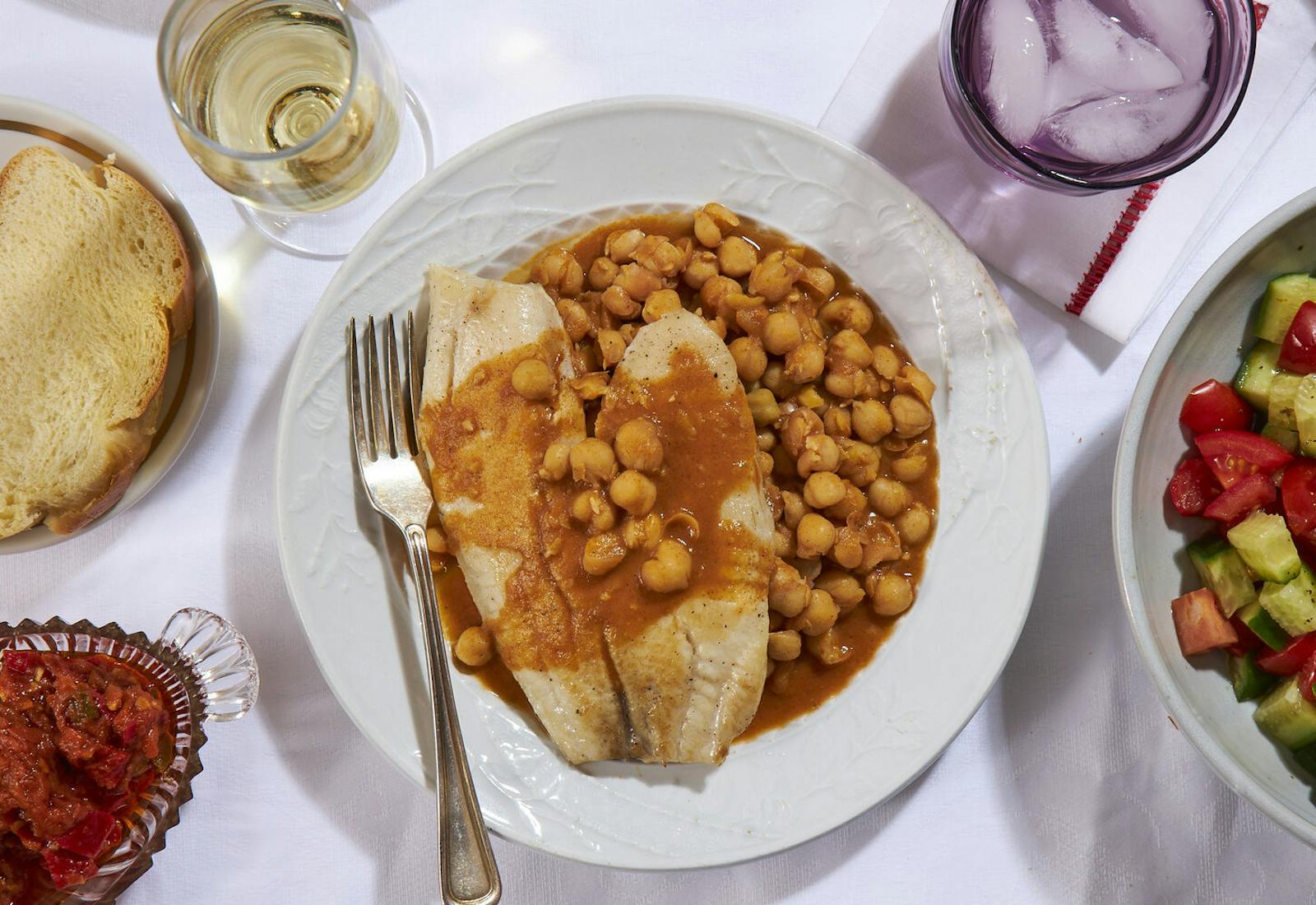 Chickpea stew with fish, slice of challah, white wine, matbucha and chopped salad atop white tablecloth.