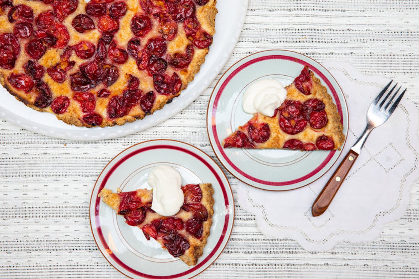 Slices of summer fruit cake with whipped cream atop grey and white tablecloth.