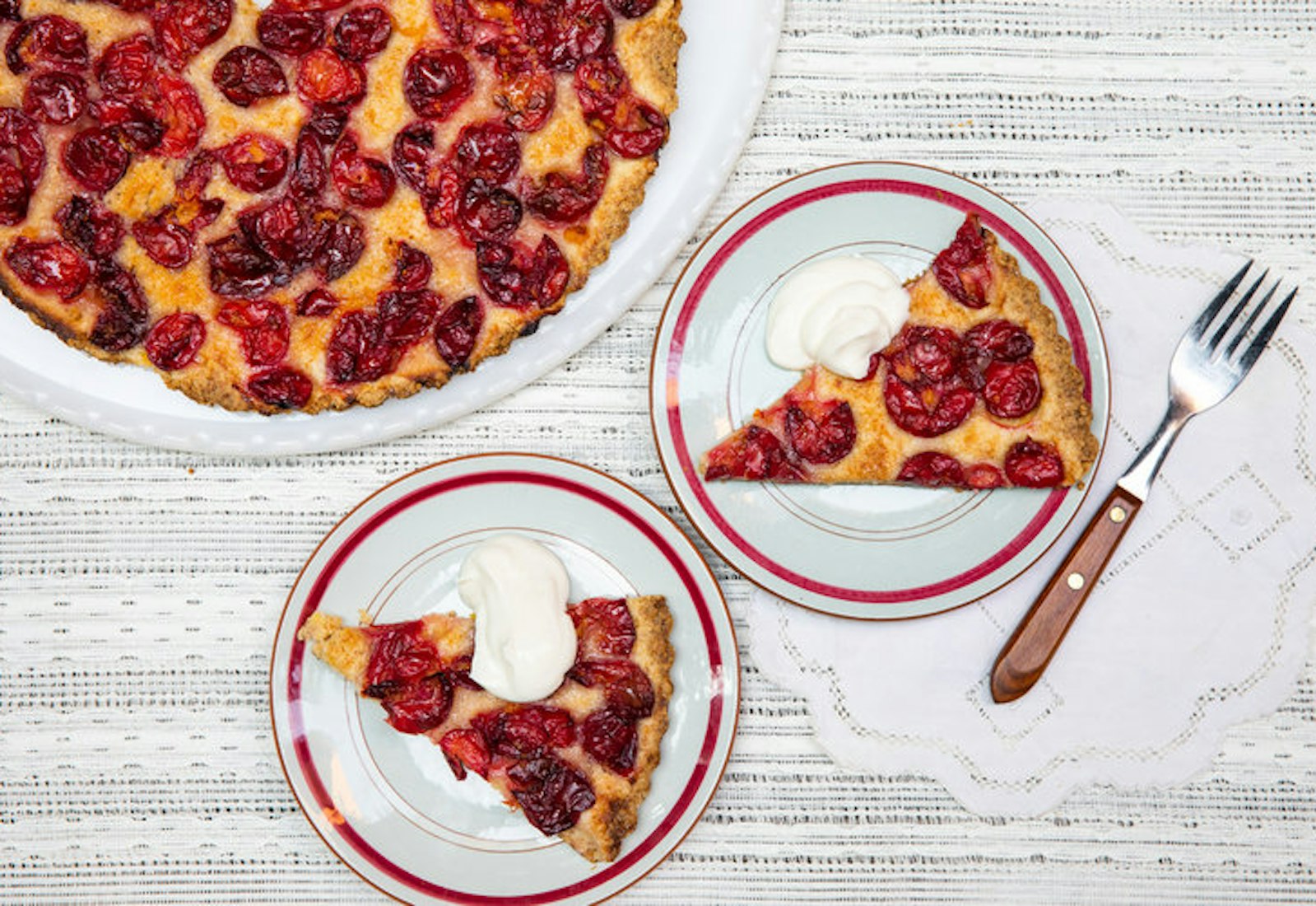 Slices of summer fruit cake with whipped cream atop grey and white tablecloth.