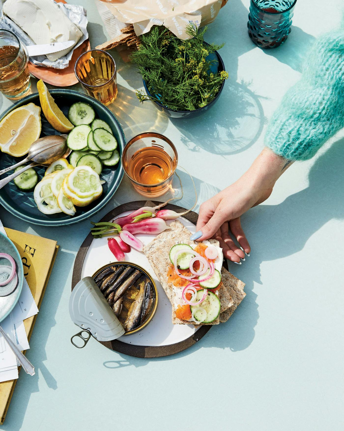 Casual table setting of crackers with cream cheese, gravlax, cucumber and red onion alongside radishes and sardines atop blue surface.