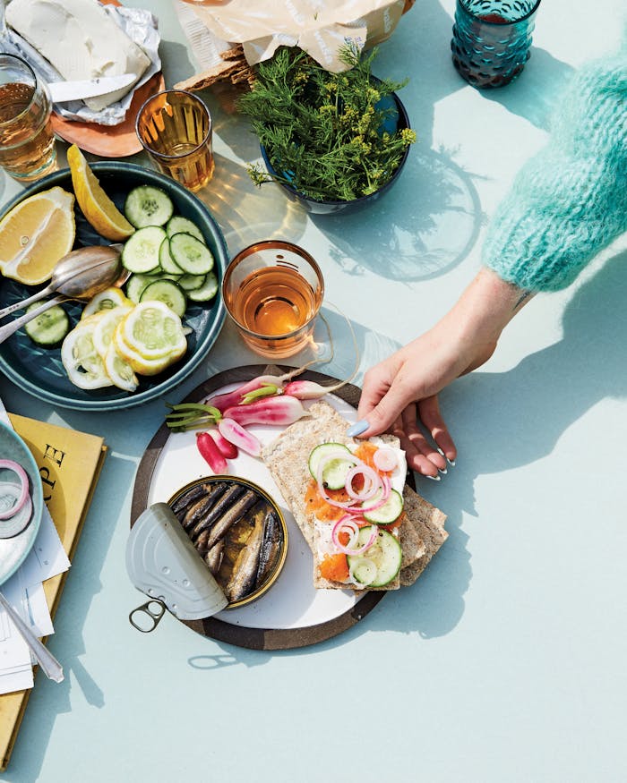 Casual table setting of crackers with cream cheese, gravlax, cucumber and red onion alongside radishes and sardines atop blue surface.