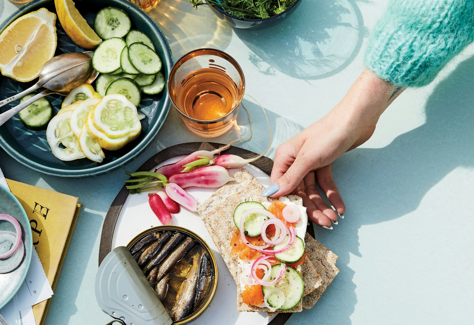 Casual table setting of crackers with cream cheese, gravlax, cucumber and red onion alongside radishes and sardines atop blue surface.