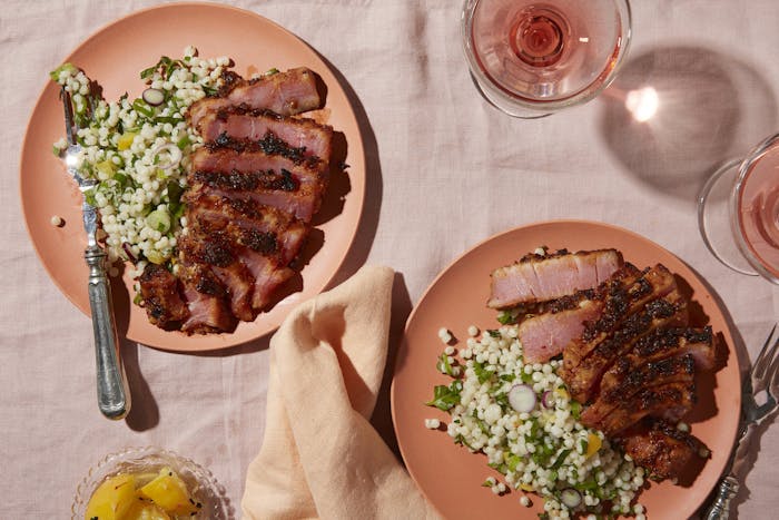 Salmon colored plates with sliced tuna pastrami and couscous salad alongside glasses of rosé, atop pink tablecloth.