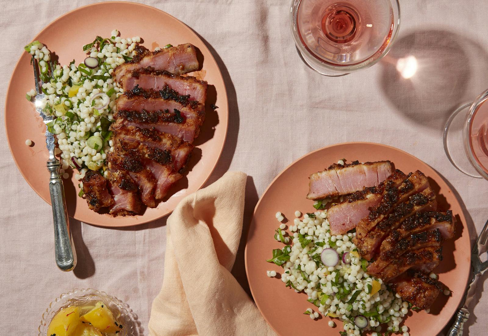 Salmon colored plates with sliced tuna pastrami and couscous salad alongside glasses of rosé, atop pink tablecloth.