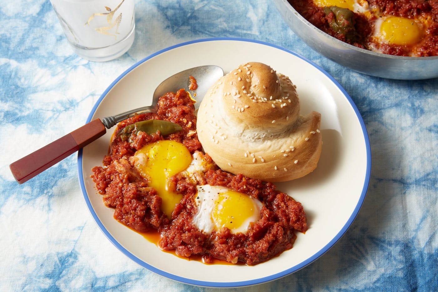 Libyan shakshuka on blue-rimmed plate with sesame seed roll atop blue tie-dyed tablecloth.