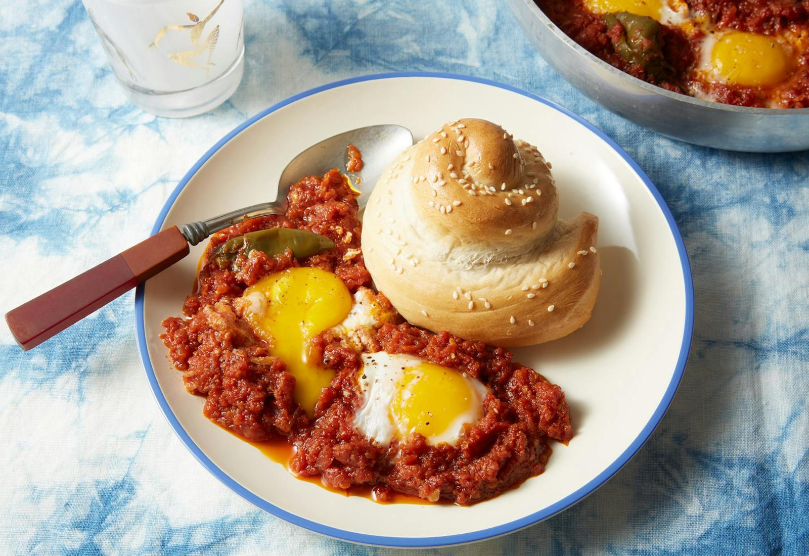 Libyan shakshuka on blue-rimmed plate with sesame seed roll atop blue tie-dyed tablecloth.