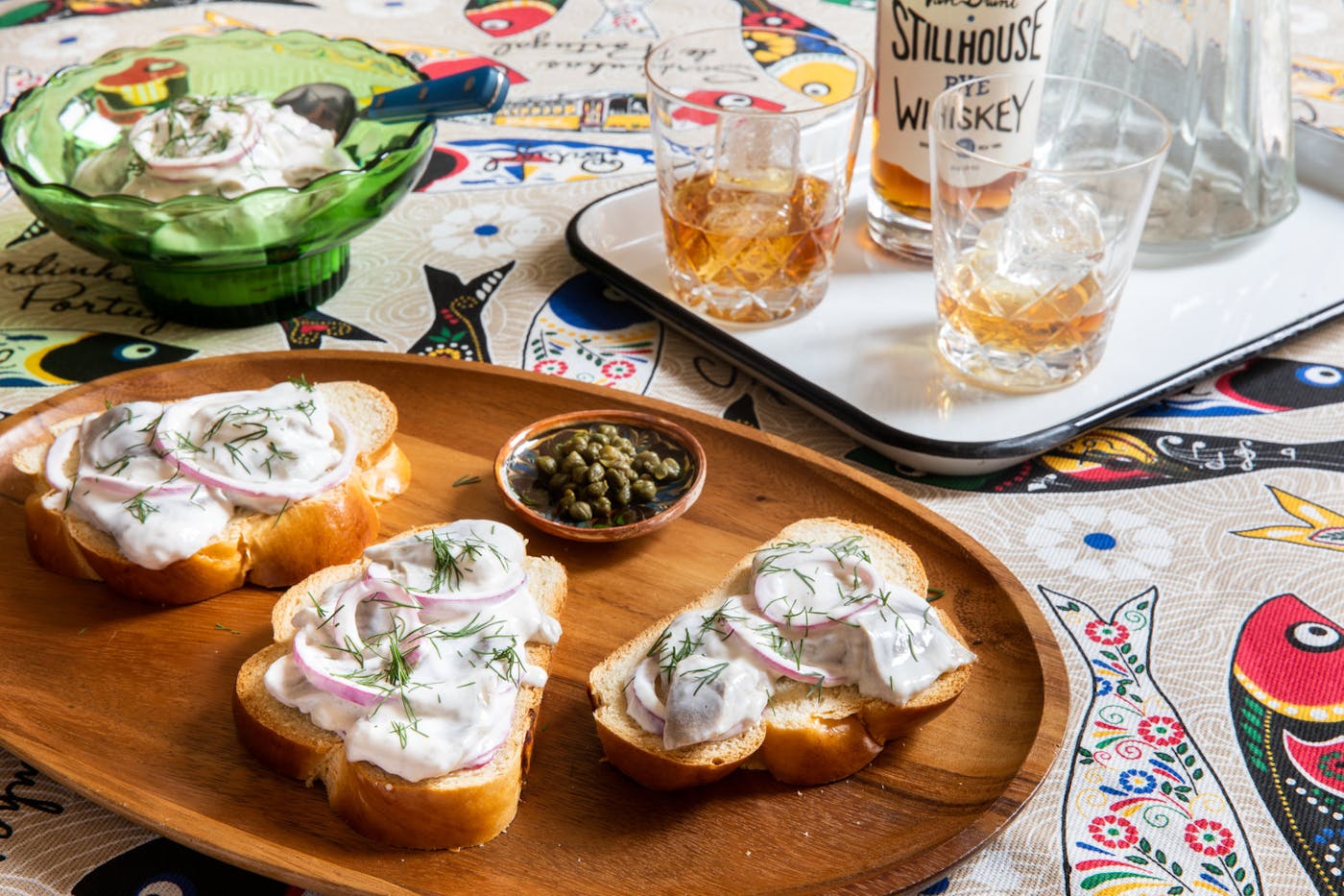 Herring in sour cream spread onto sliced challah garnished with red onion and dill alongside dish of capers and tray of whiskey glasses, atop fish-print tablecloth.