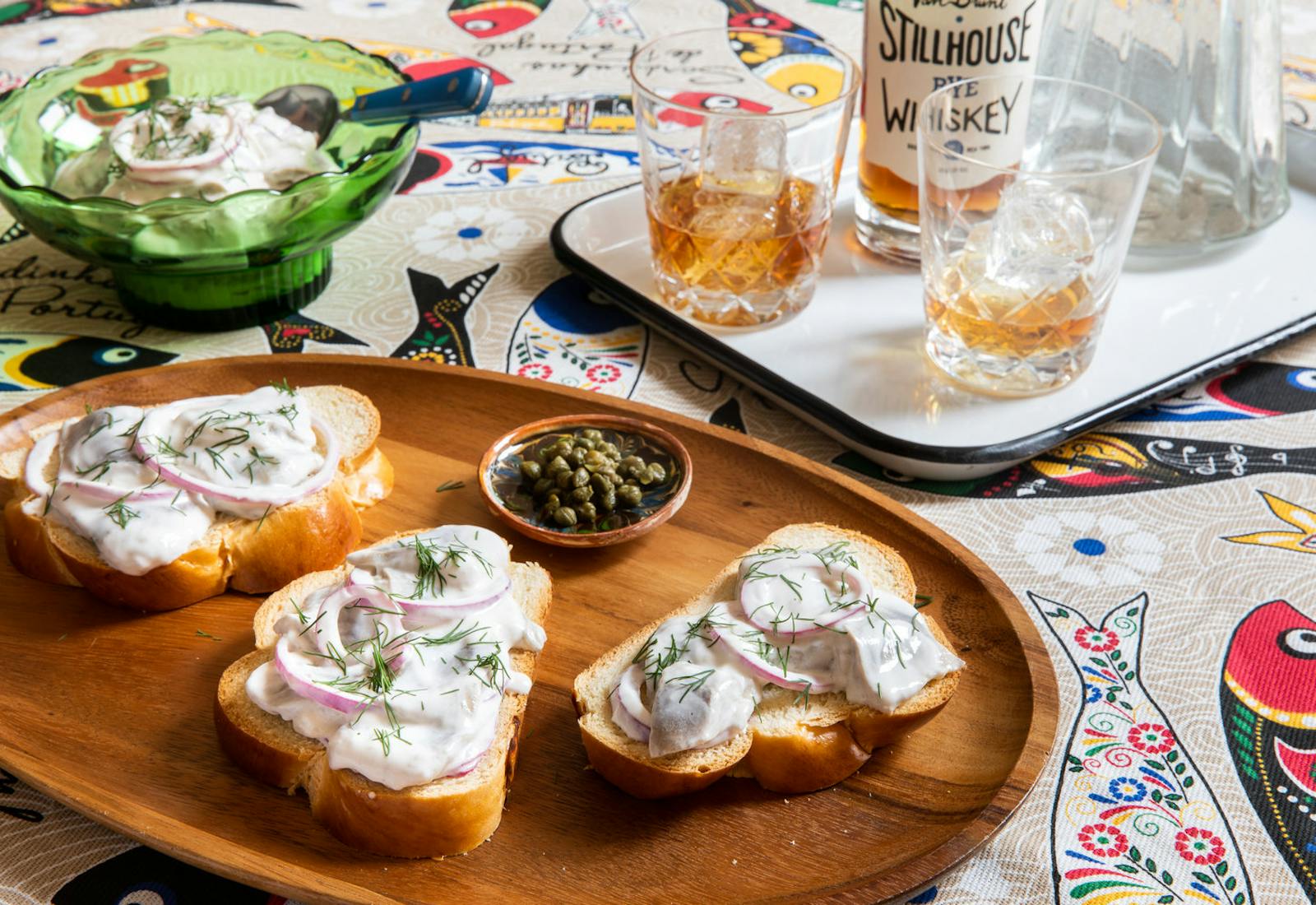 Herring in sour cream spread onto sliced challah garnished with red onion and dill alongside dish of capers and tray of whiskey glasses, atop fish-print tablecloth.