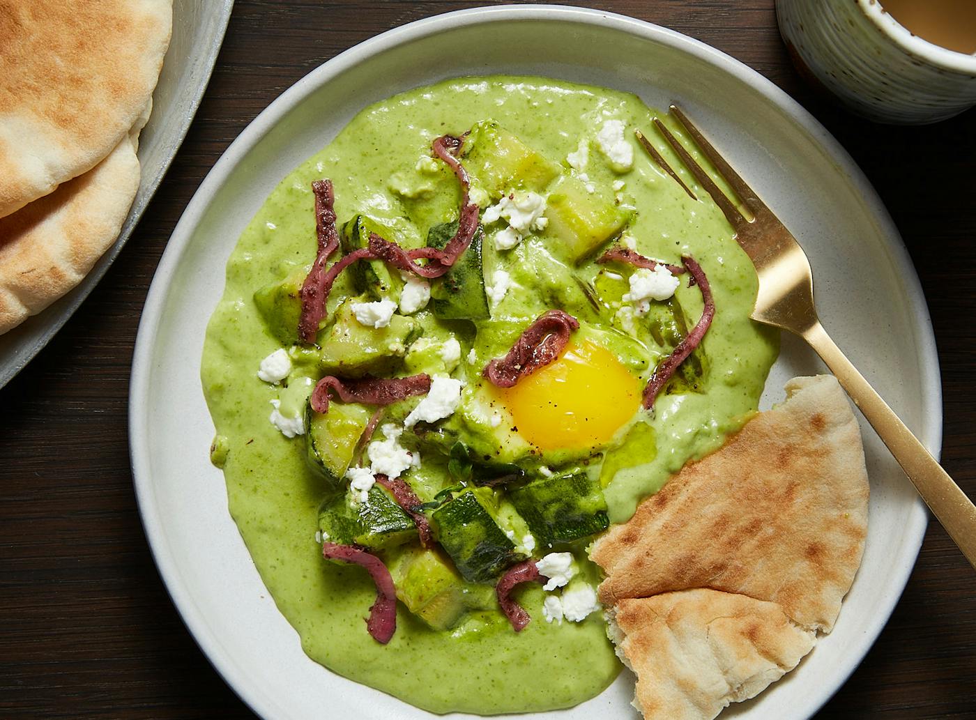 Green shakshuka with crumbled goat cheese and pickled onions accompanied by fresh pita, atop wooden table.