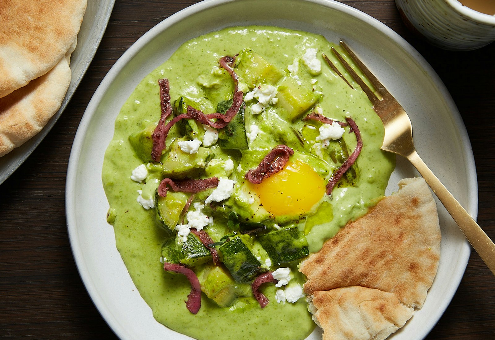 Green shakshuka with crumbled goat cheese and pickled onions accompanied by fresh pita, atop wooden table.