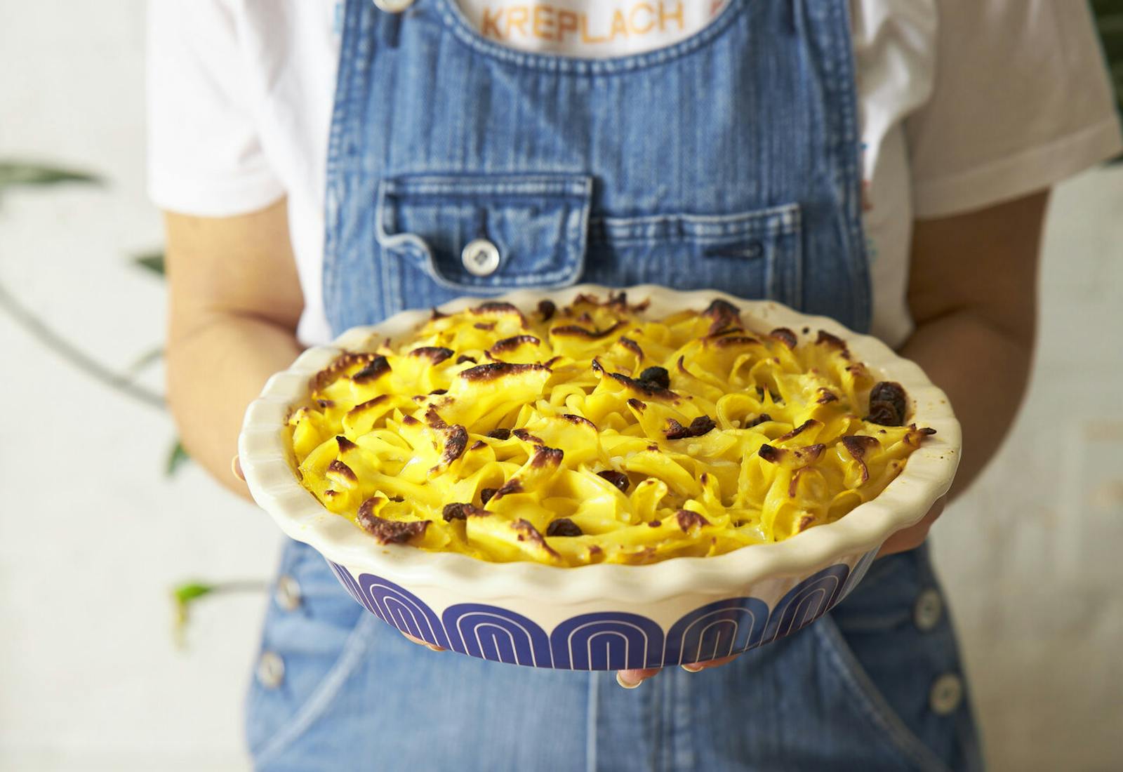 Chef in denim apron holding sweet noodle kugel in blue and white casserole dish.