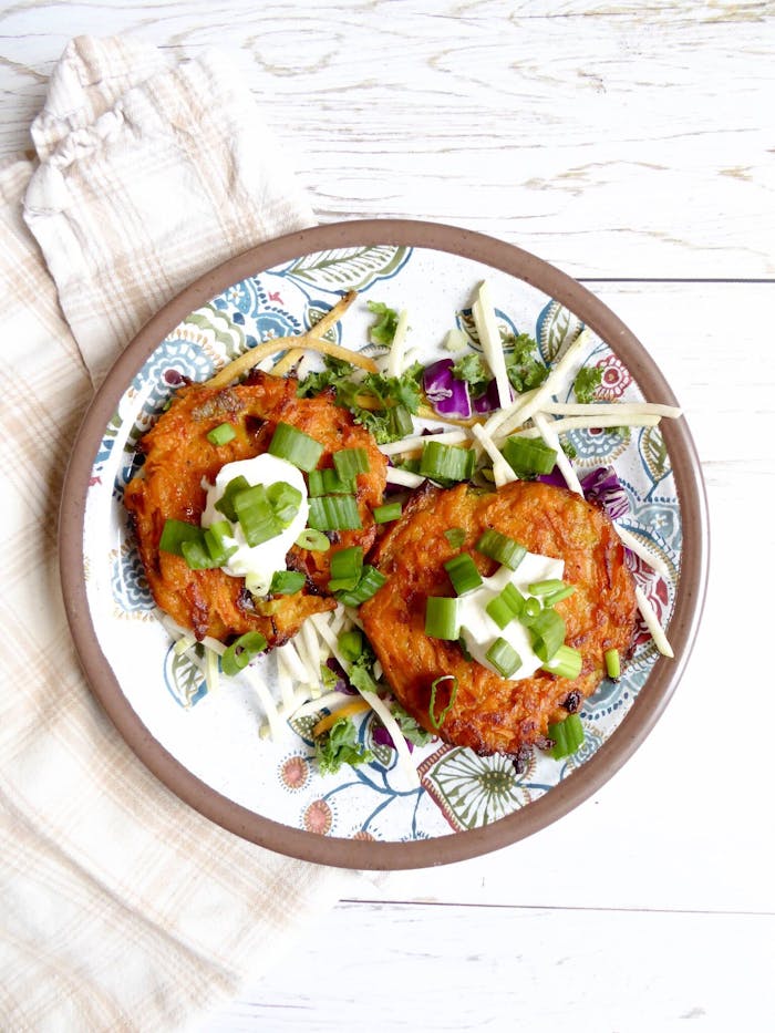 Two sweet potato latkes with dollops of sour cream and scallions with cabbage salad atop white wooden table.