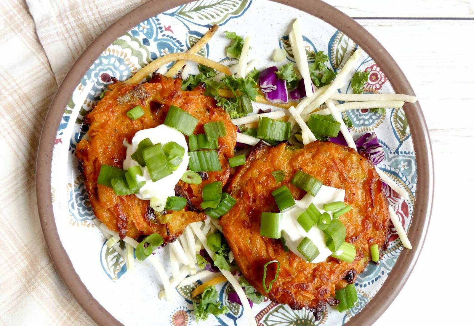 Two sweet potato latkes with dollops of sour cream and scallions with cabbage salad atop white wooden table.